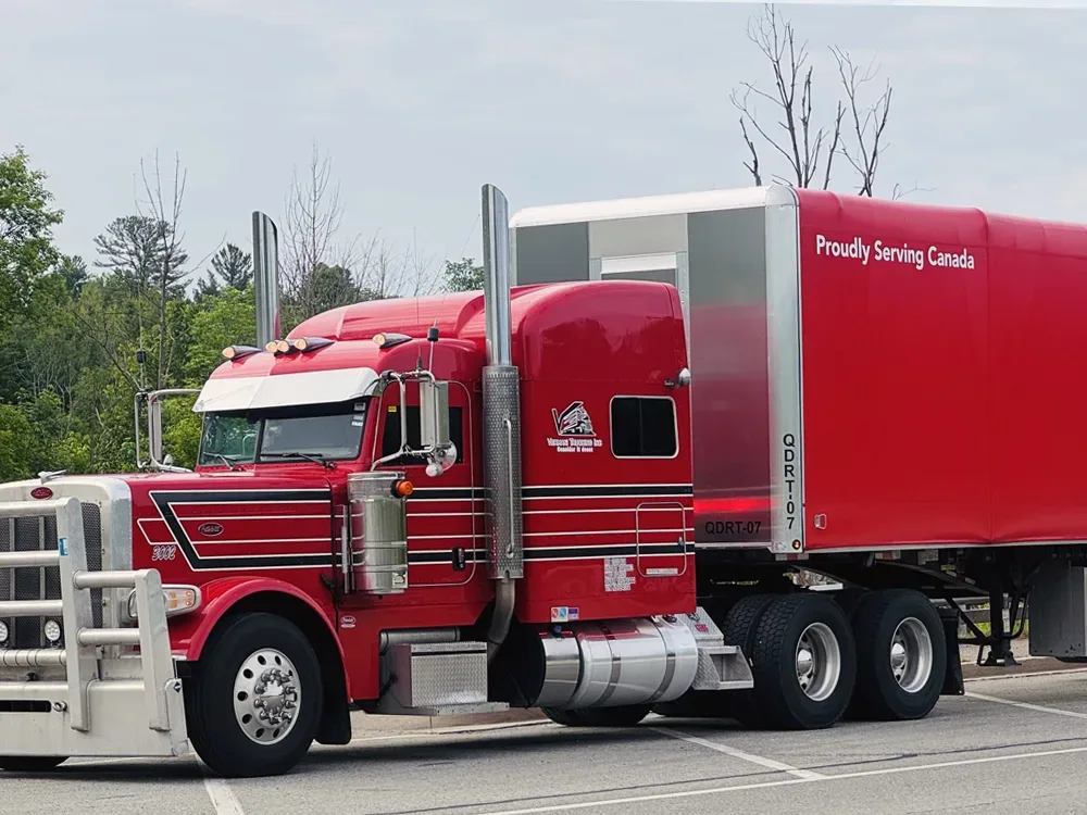 Camion rouge de transport avec l'inscription "Proudly Serving Canada" stationné dans un parking, entouré d'arbres.