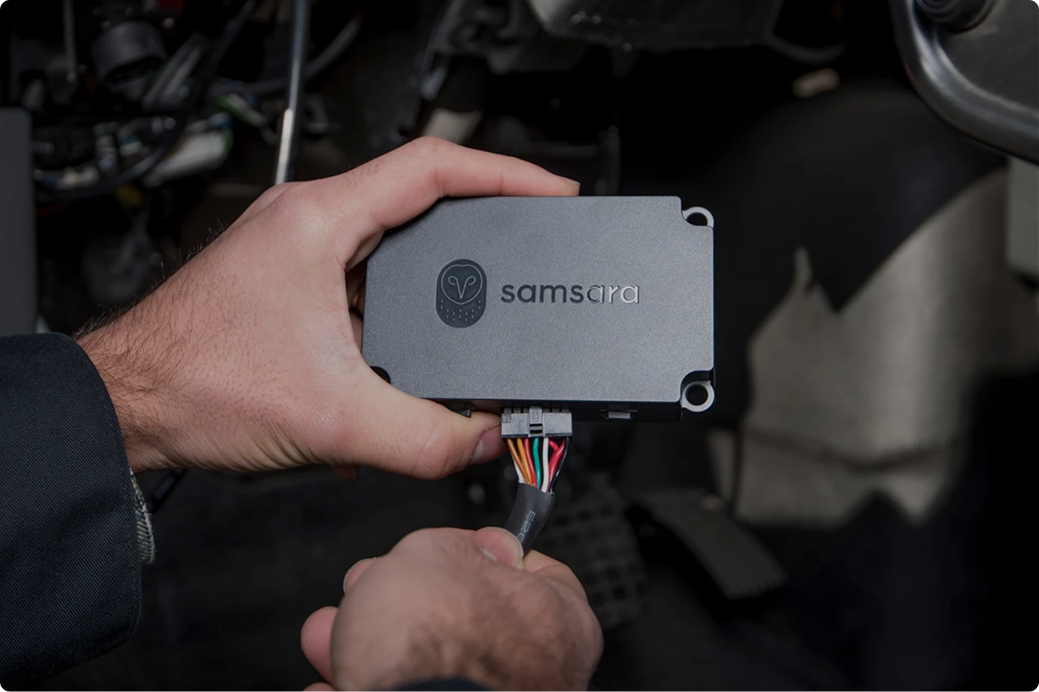 Una mano conectando un dispositivo Samsara gris con cables de colores dentro del compartimiento del motor de un vehículo.