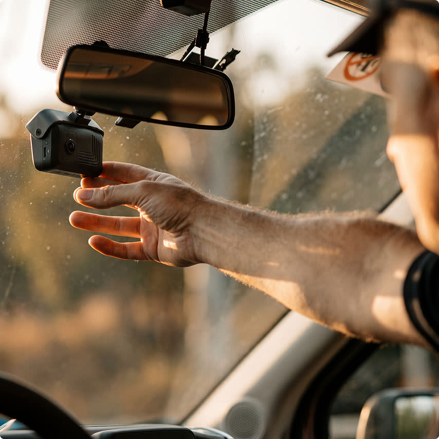 A person's hand reaching to adjust a black dash cam mounted near the rearview mirror inside a car.