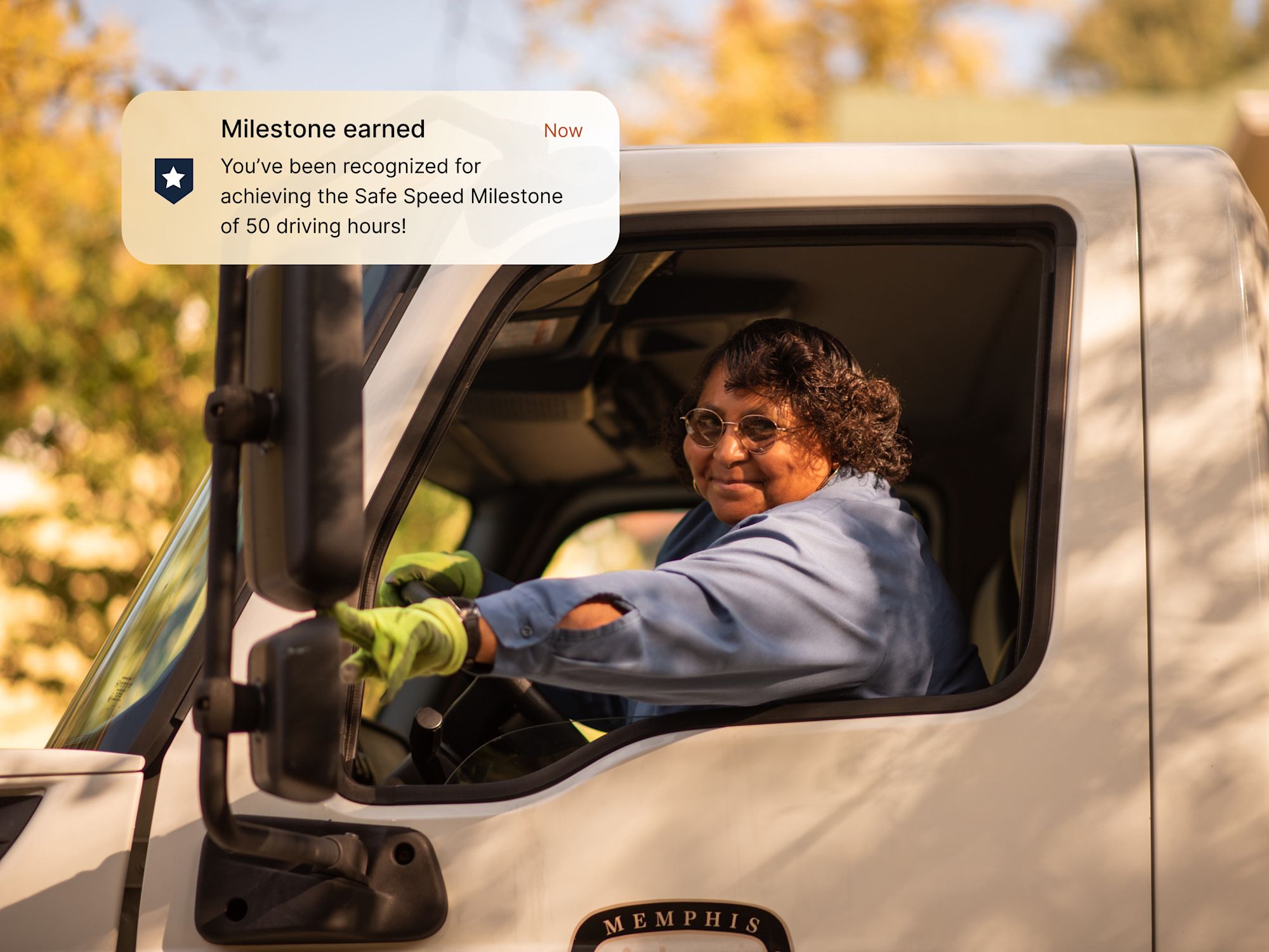 A smiling female truck driver in a white cab with a milestone notification for achieving 50 safe driving hours.