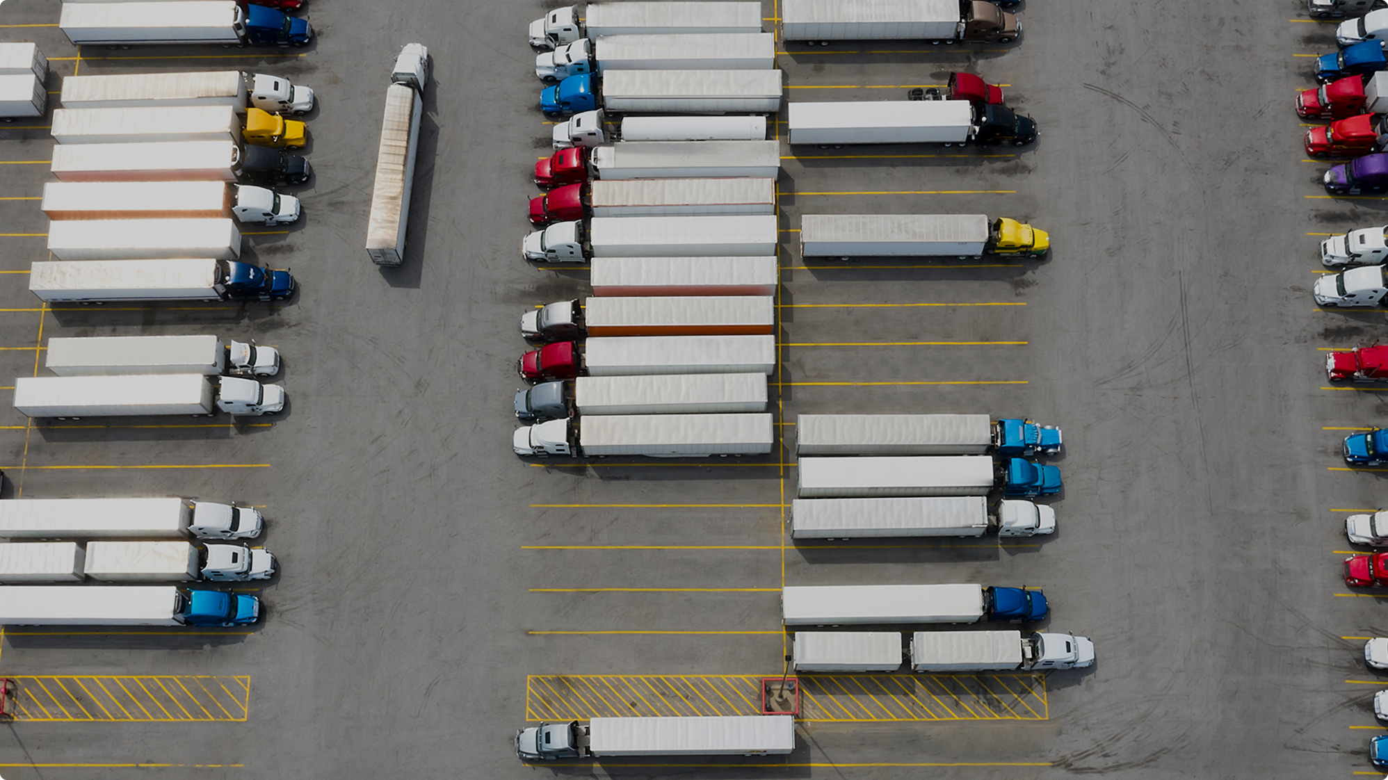 Aerial view of a truck depot with colorful semi-trucks and white trailers parked in organized rows on a concrete lot.