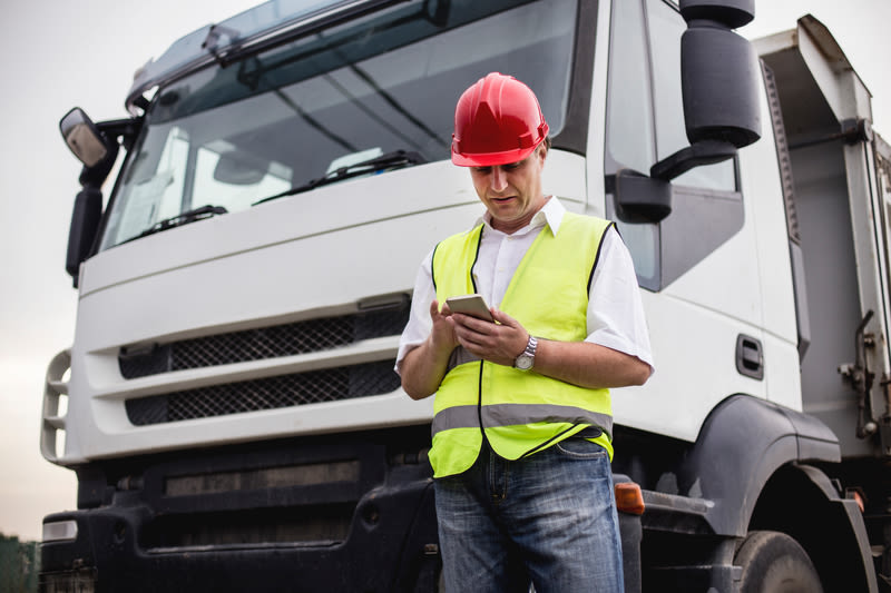 Man met rode helm en geel veiligheidsvest bekijkt smartphone voor een witte vrachtwagen.