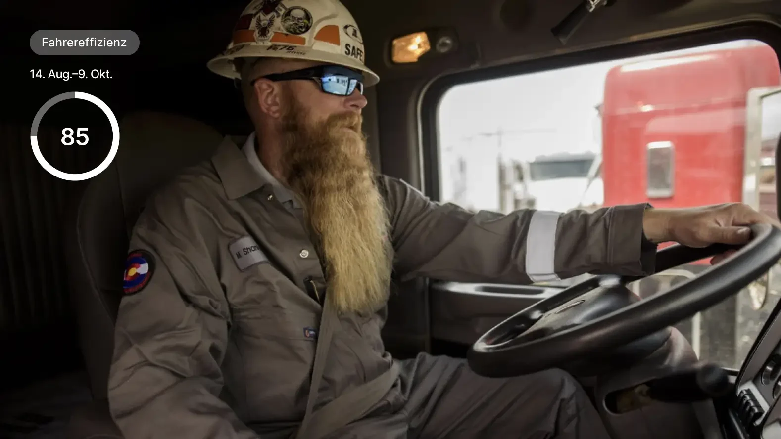 Bauarbeiter mit Bart fährt LKW und trägt Schutzhelm, auf dem eine Fahrer-Effizienzbewertung von 85 angezeigt wird.