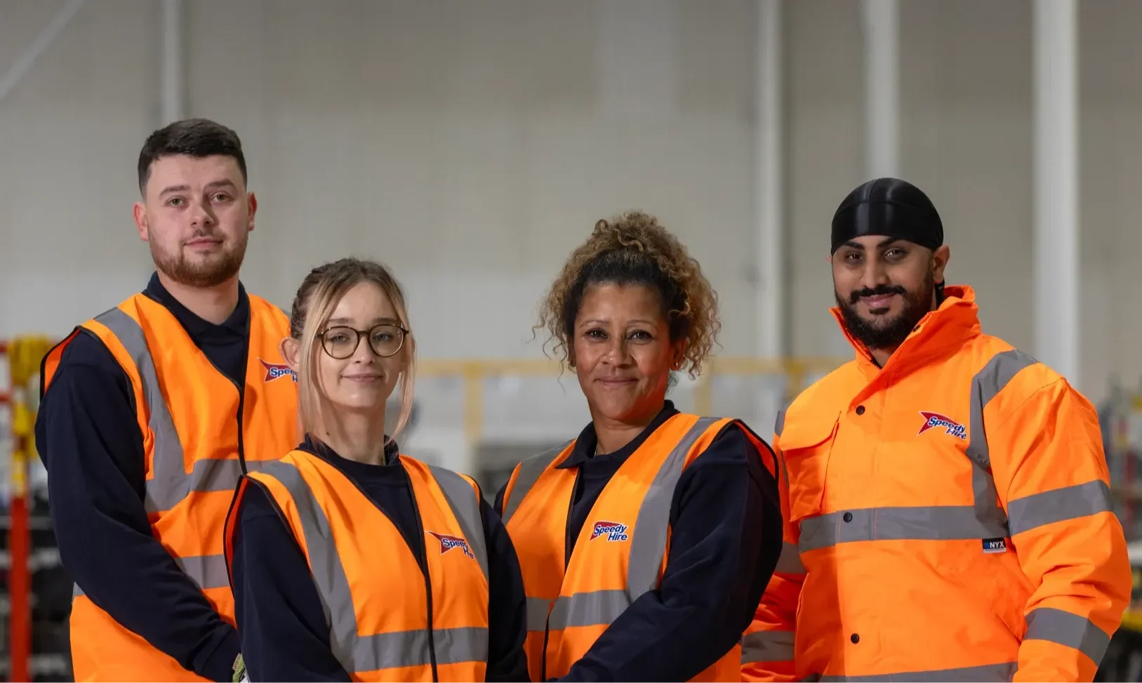 Four Speedy Hire employees in orange high-visibility vests stand together in a large warehouse, smiling at the camera.
