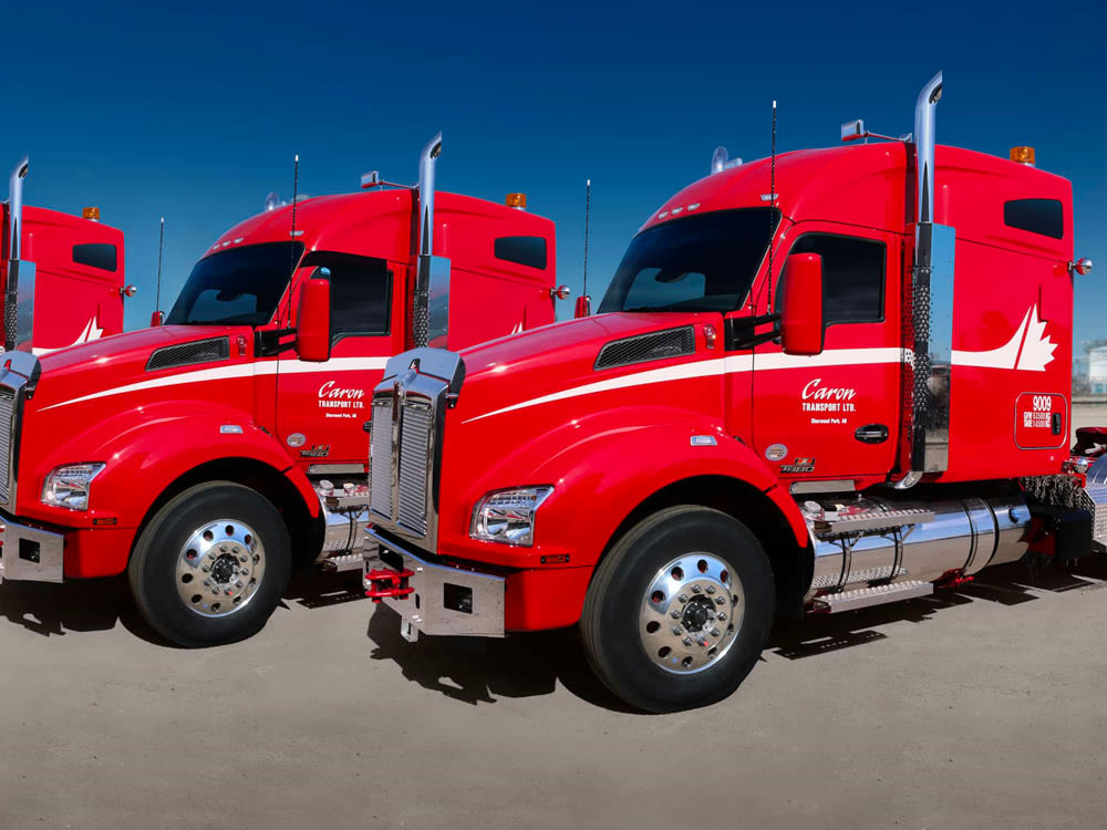 Rangée de semi-remorques Caron Transport rouge vif avec des accents blancs, garés côte à côte dans un ciel bleu clair.