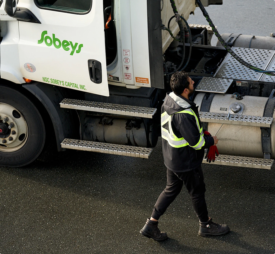 Worker in safety vest walking beside a white Sobeys delivery truck, holding red gloves.