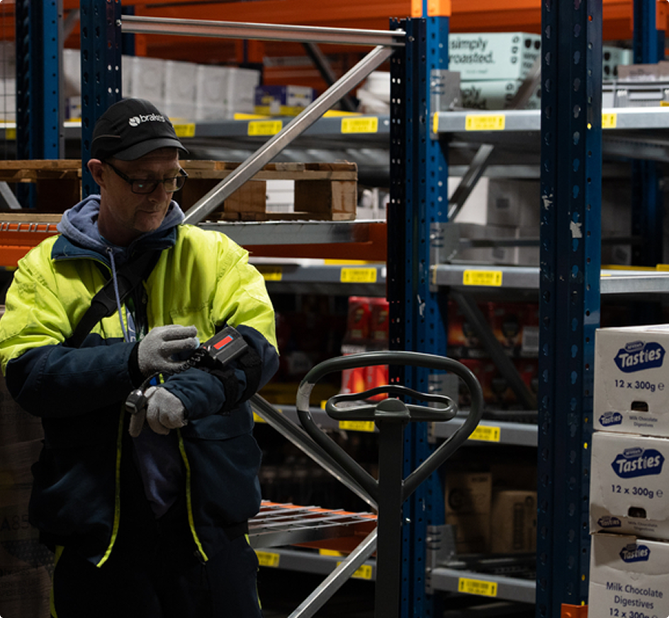 Magazijnmedewerker in fluorescerende jas gebruikt een scanner bij een palletwagen tussen stellingen met voedselproducten.