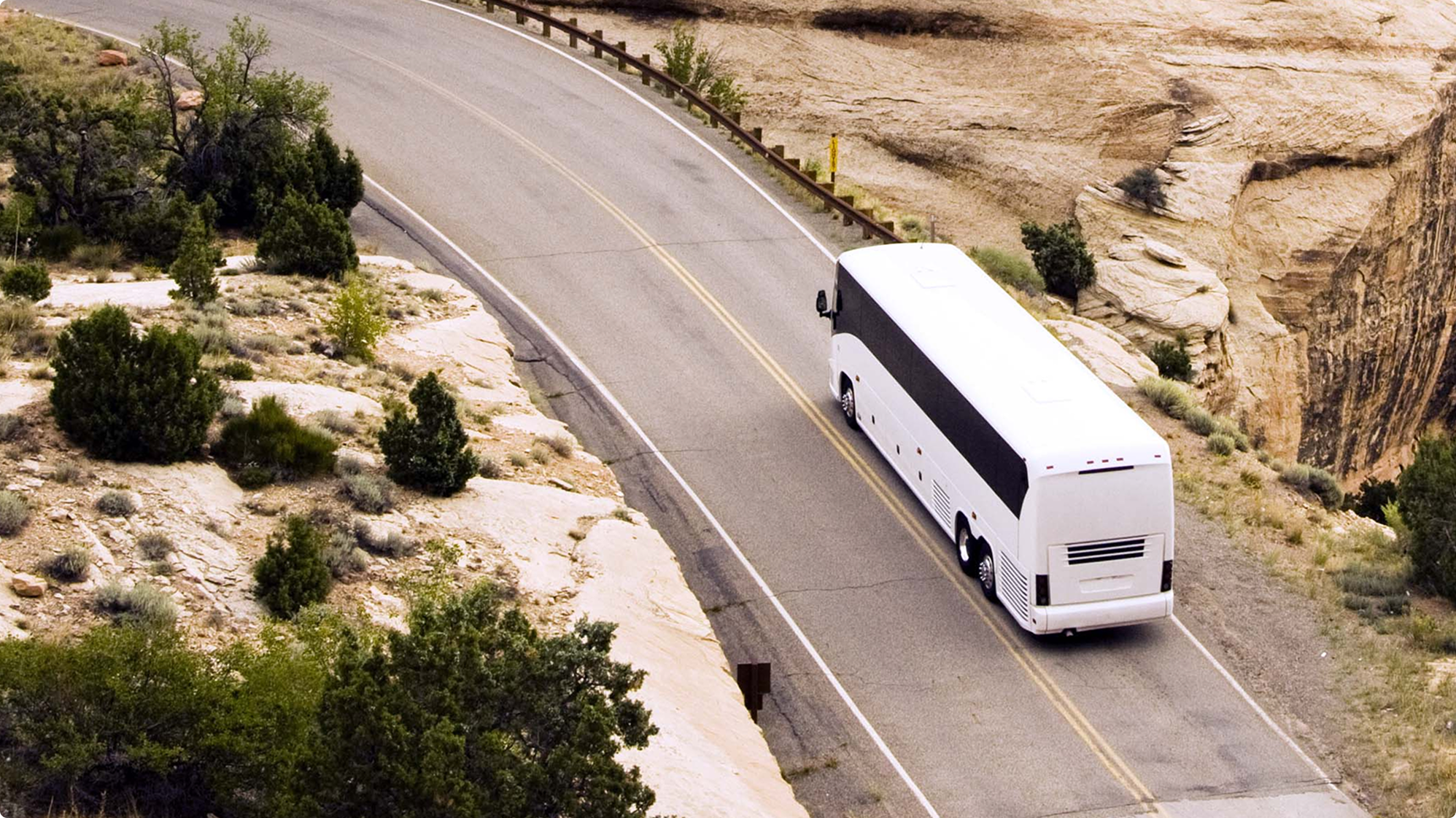 White tour bus driving along a winding desert highway with rocky terrain and sparse vegetation on both sides.
