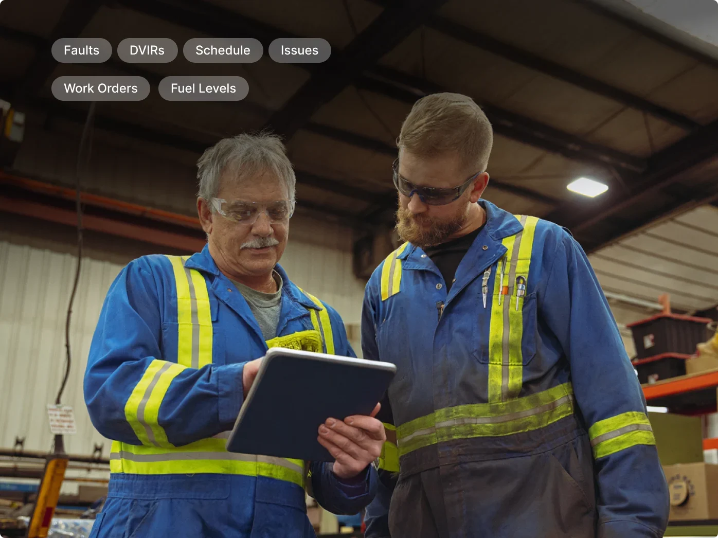 Two industrial workers in blue safety uniforms with reflective stripes reviewing information on a tablet in a warehouse.