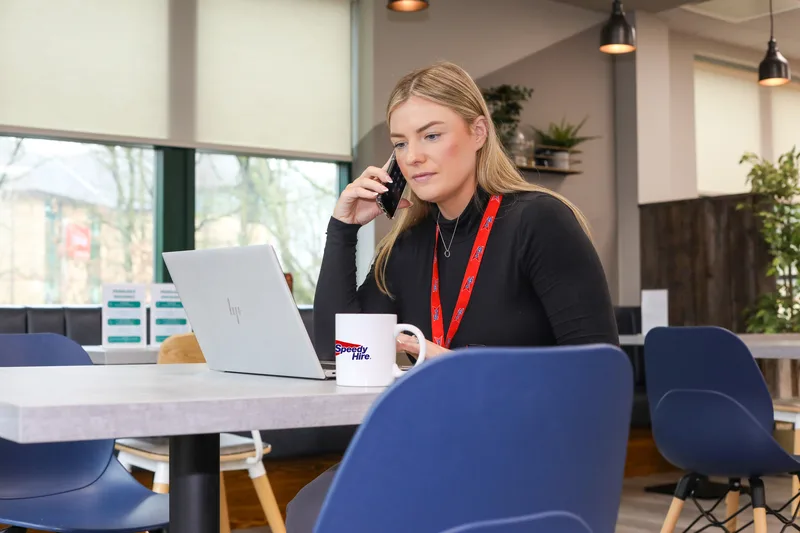 A woman in a black top talks on a phone at a laptop, with a Speedy Hire mug on the desk in a modern office.