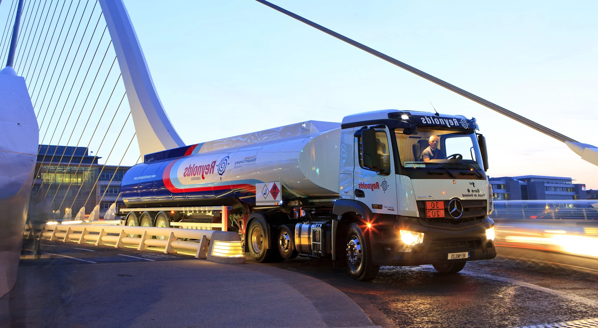 Een Reynolds tankwagen rijdt over een moderne brug met kabels bij schemering, de koplampen verlichten de weg.