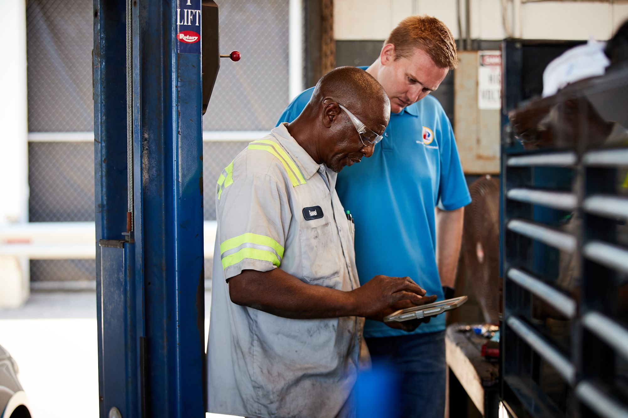 Two mechanics in an auto shop examining a tablet, one in a gray uniform with safety stripes and one in blue.