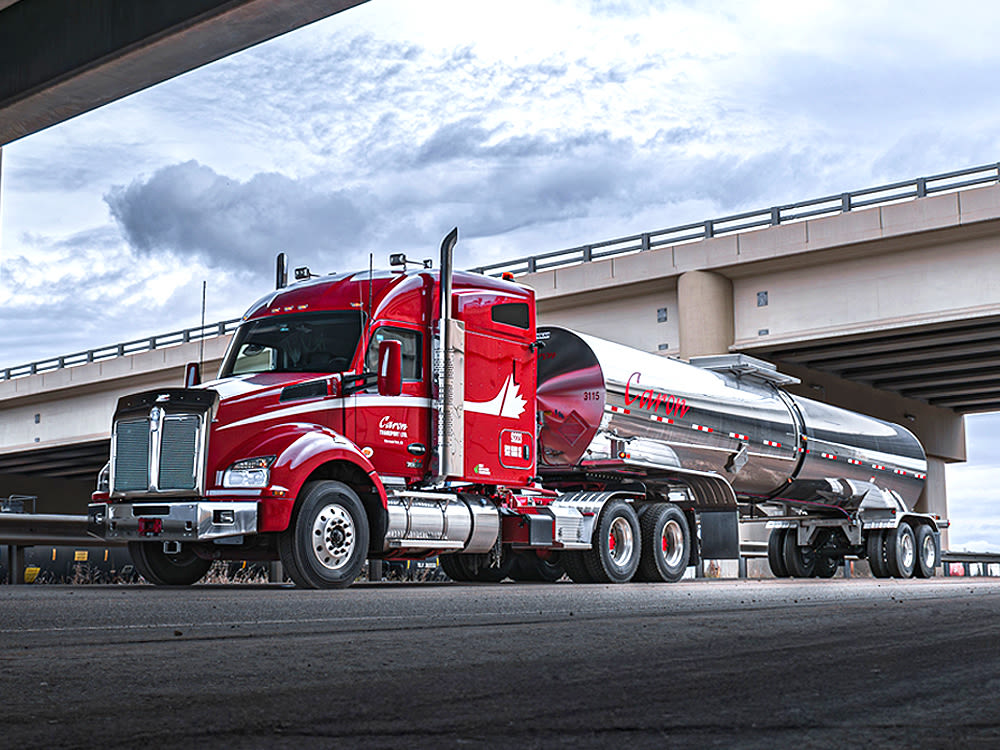 Camion Kenworth rouge avec remorque citerne chromée garé sous un pont autoroutier, sous un ciel nuageux.