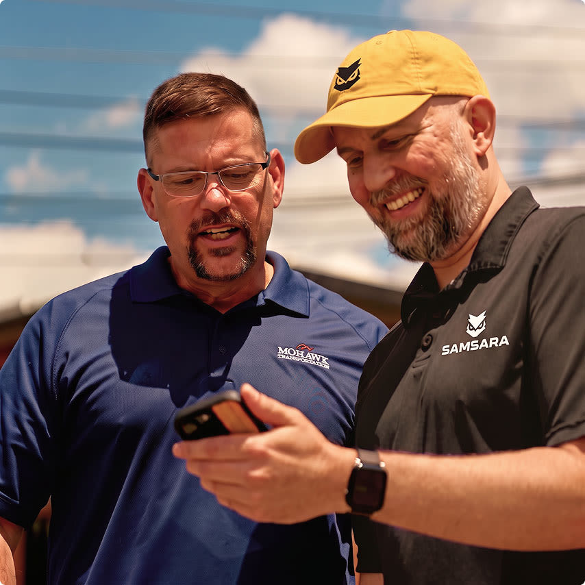 Two men, one in a Mohawk Transportation shirt and one in a Samsara shirt, smile while looking at a smartphone outdoors.