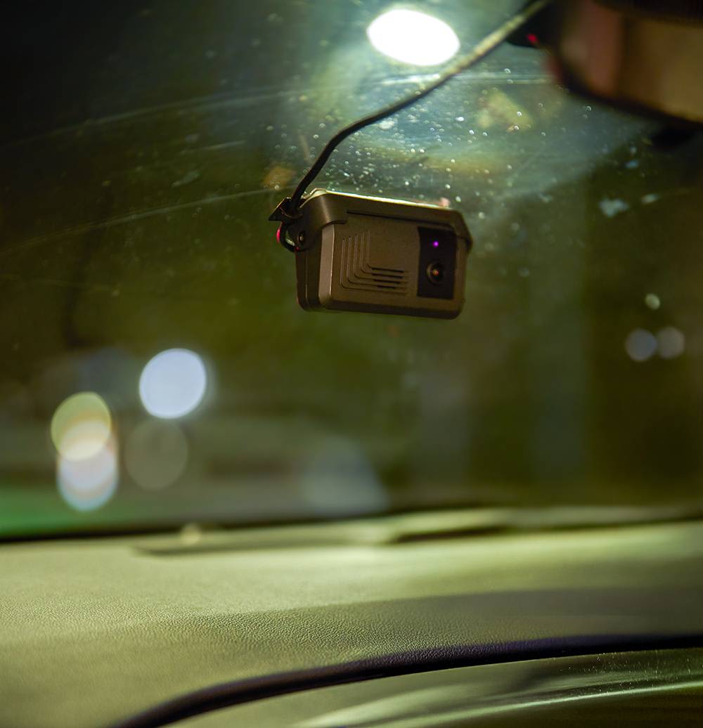 Small dashboard camera mounted on car windshield at night with city lights blurred in the background.
