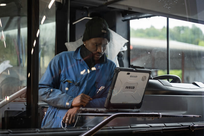 A technician in a blue jumpsuit and black beanie uses an Iveco rugged laptop inside a bus cabin.