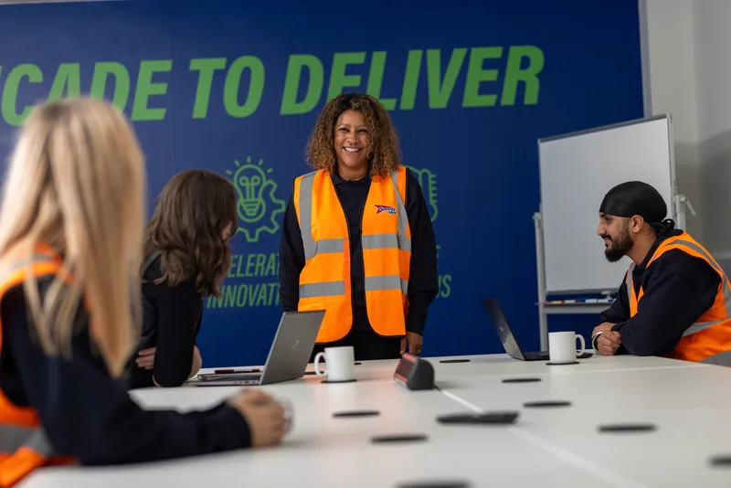 A smiling woman in an orange safety vest leads a meeting with colleagues at a conference table in front of a blue wall.