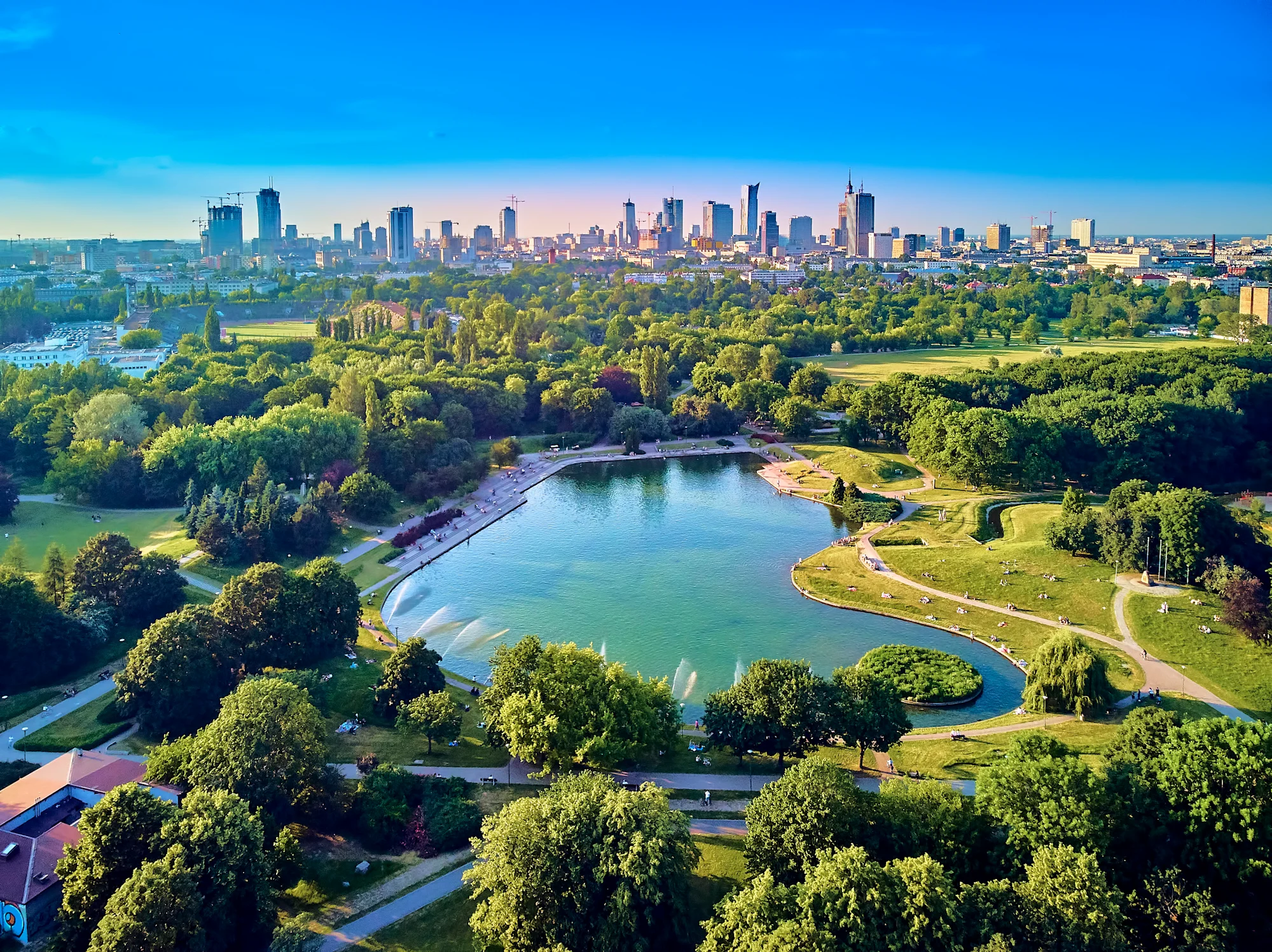 Aerial view of a city park with a blue lake surrounded by green trees, with a modern skyline visible in the background.