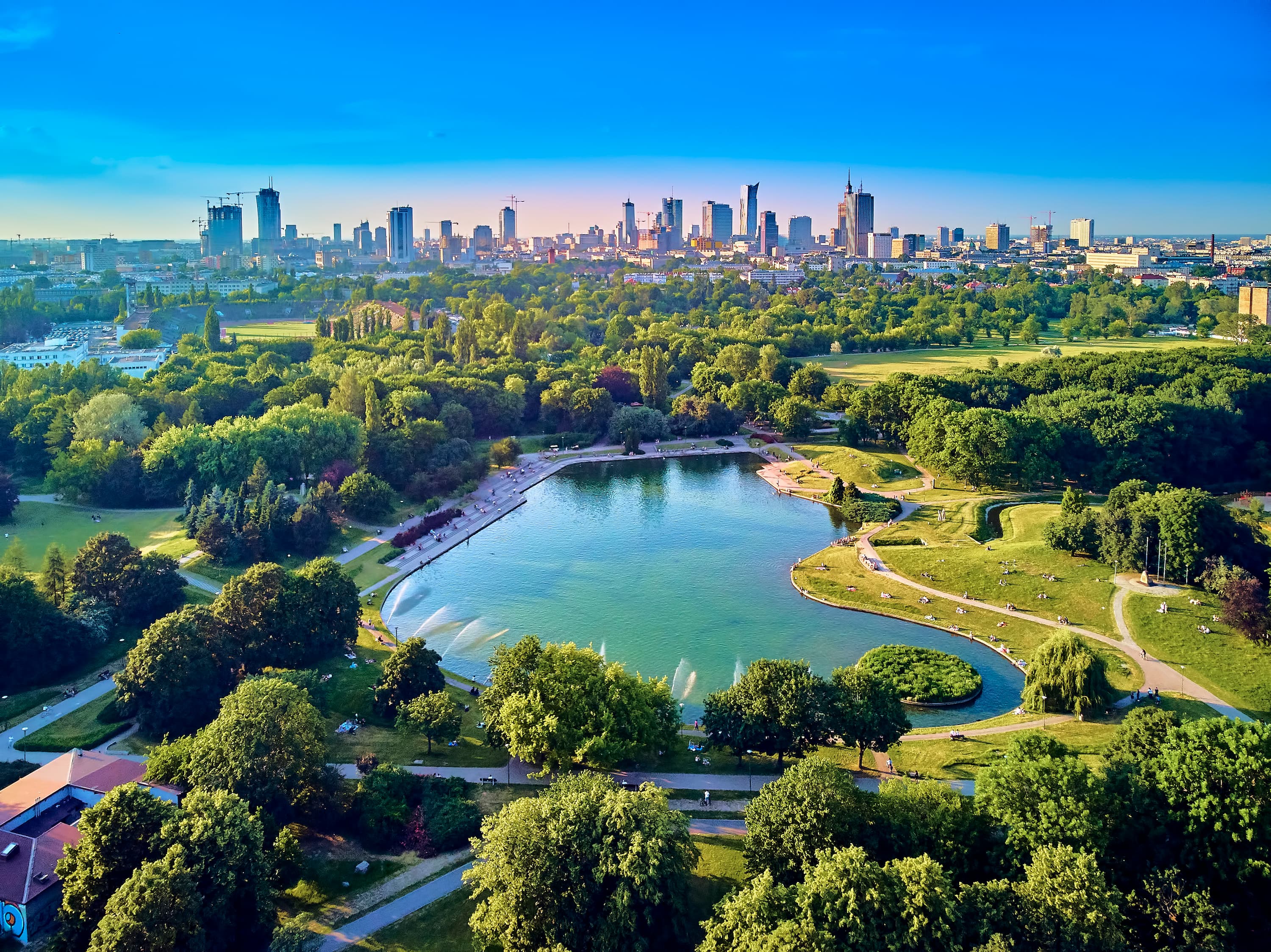 Aerial view of a city park with a blue lake surrounded by green trees, with a modern skyline visible in the background.