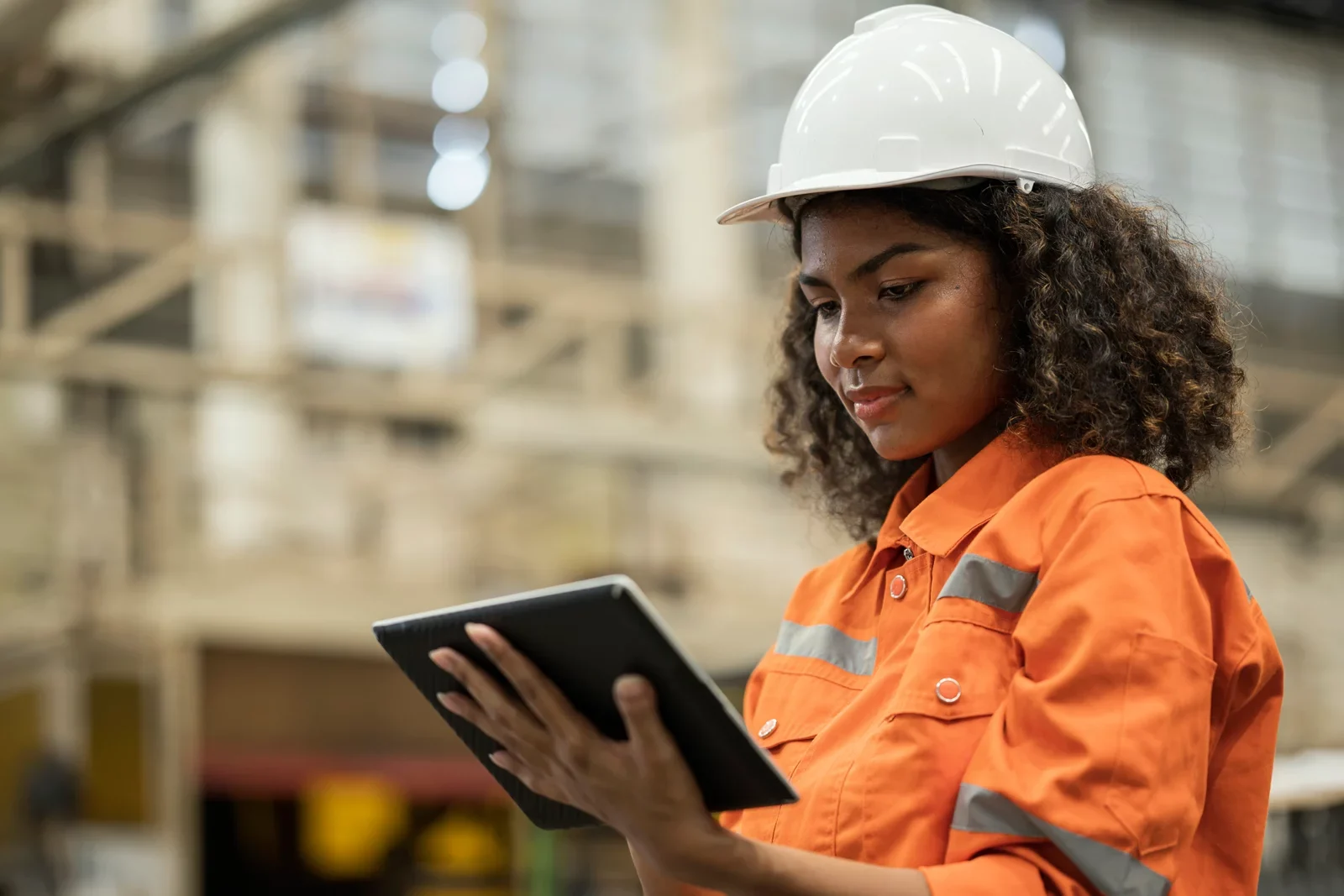 A female engineer in an orange hi-vis suit and white hard hat reviews data on a tablet at an industrial site.