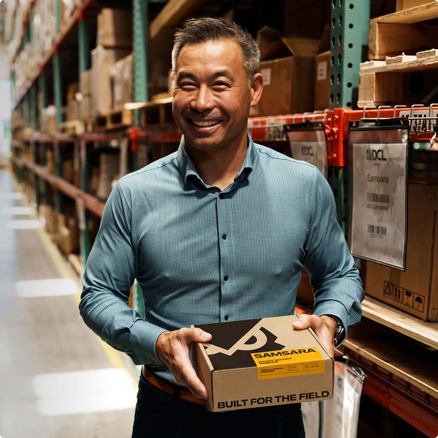 Smiling man in a blue shirt holds a Samsara branded box in a warehouse with shelves of cardboard boxes.