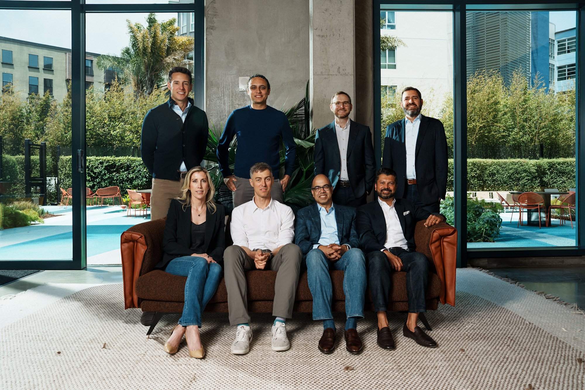 Eight professionals pose for a group photo indoors, with four seated on a brown sofa and four standing behind them.