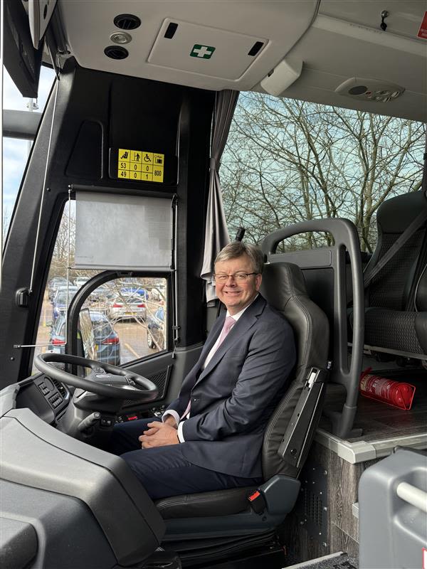 A smiling man in a dark suit sits in the driver's seat of a modern coach, with a steering wheel and capacity signs visible.