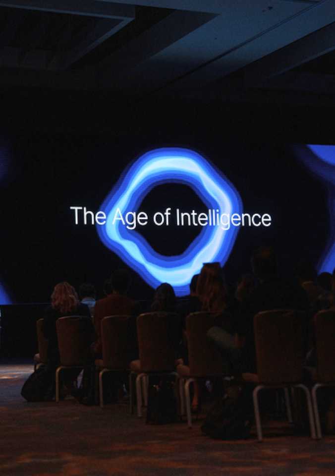 Audience watching presentation with "The Age of Intelligence" text and glowing blue circular logo on dark screen.