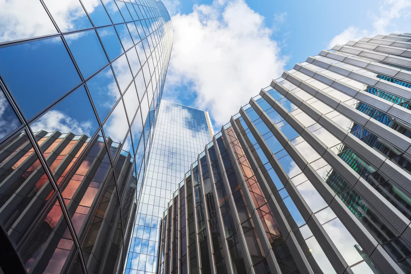 Looking up at modern glass skyscrapers reflecting a blue sky with white clouds, shot from ground level.