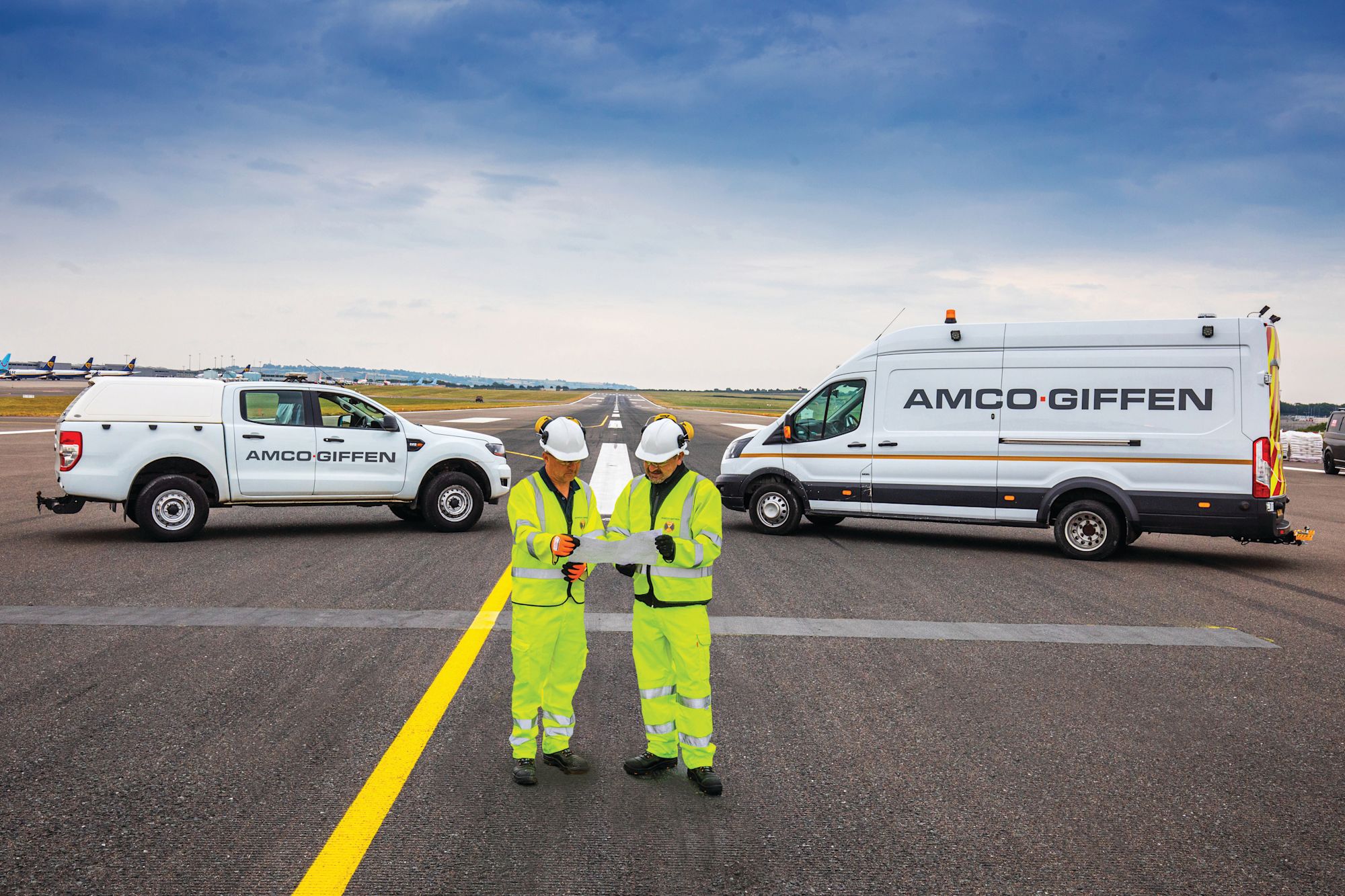 Two Amco Giffen workers in hi-vis gear review documents on an airport runway, flanked by branded white vehicles.