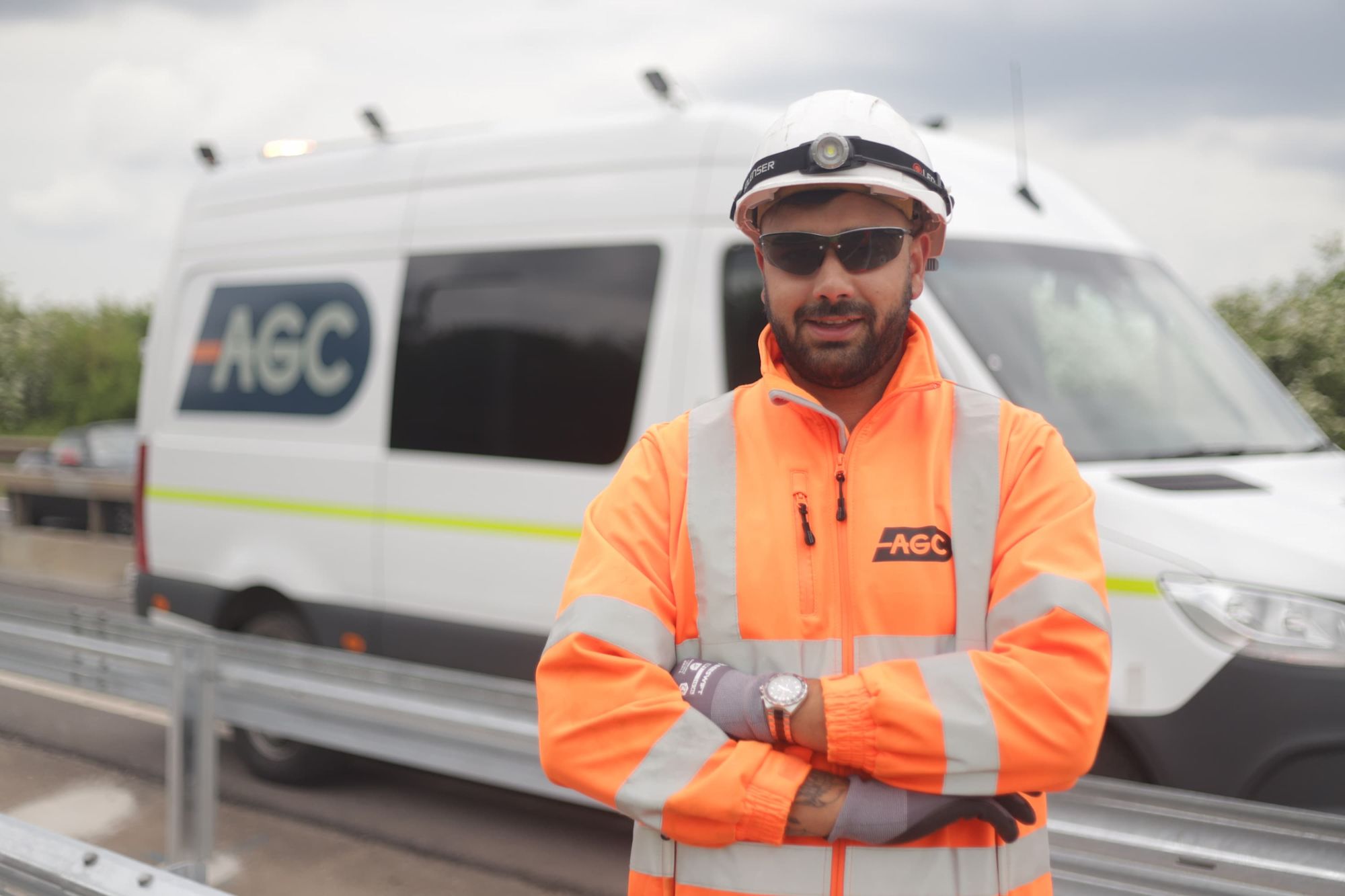 AGC worker in orange hi-vis jacket and white hard hat stands arms crossed beside an AGC van on a motorway.