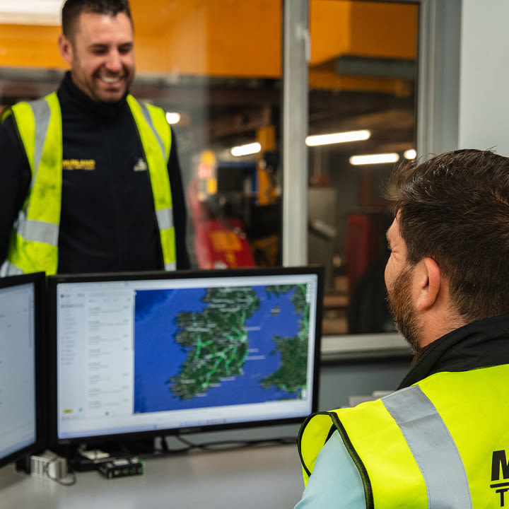 Two workers in hi-vis vests at a control desk with dual monitors displaying a map of Ireland and the UK.