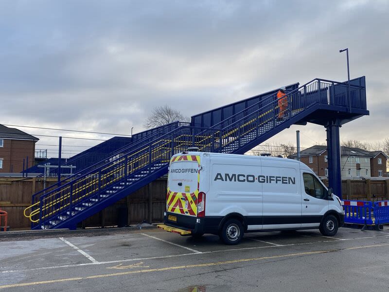 An Amco Giffen van parked beside a blue steel footbridge with yellow handrails at a railway station construction site.