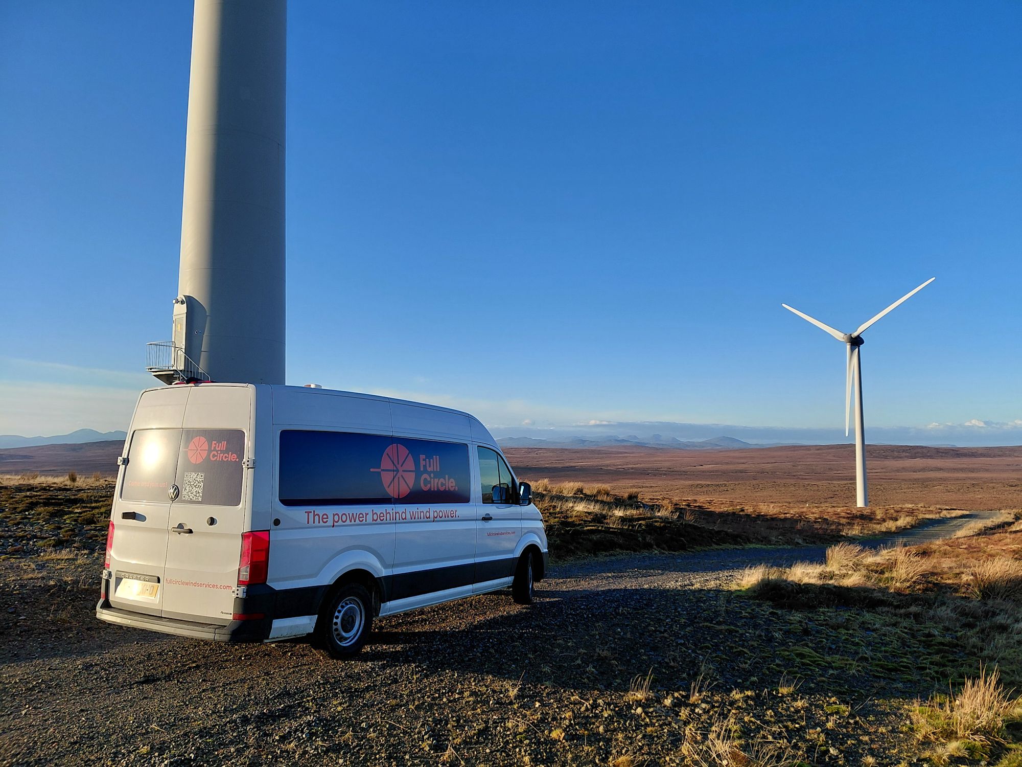 A Full Circle branded white van parked beside a wind turbine on open moorland under a clear blue sky.