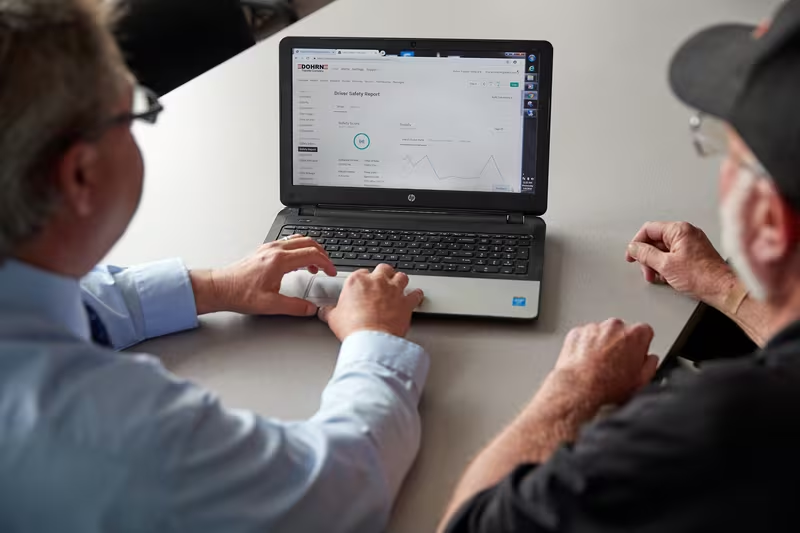 Two men reviewing a Driver Safety Report on an HP laptop, viewed from above at a table.