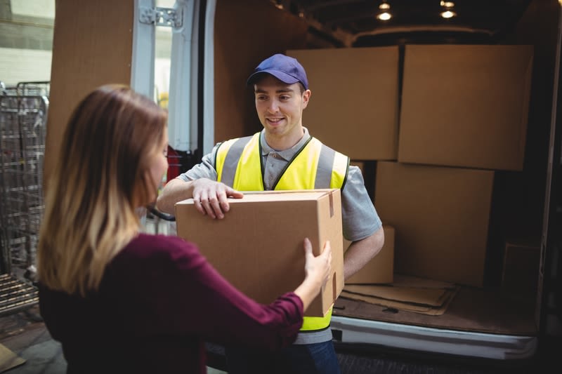 Ein Zusteller in gelber Warnweste übergibt einer Frau ein Paket vor einem geöffneten Lieferwagen.