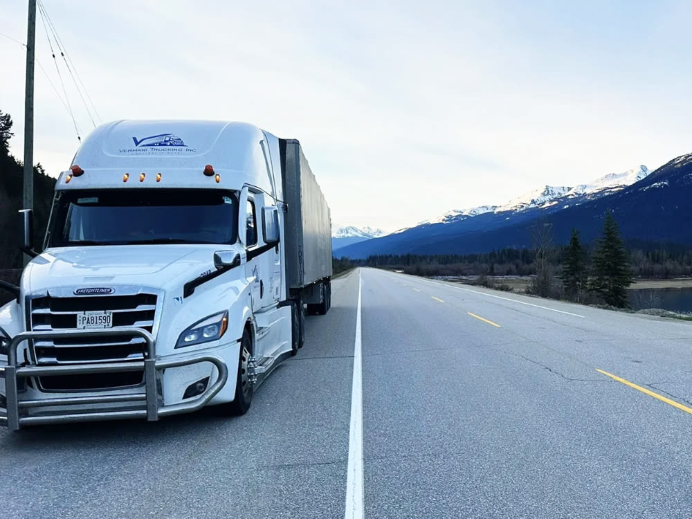 Un semi-remorque blanc circule sur une autoroute avec des montagnes enneigées en arrière-plan, sous un ciel bleu clair.
