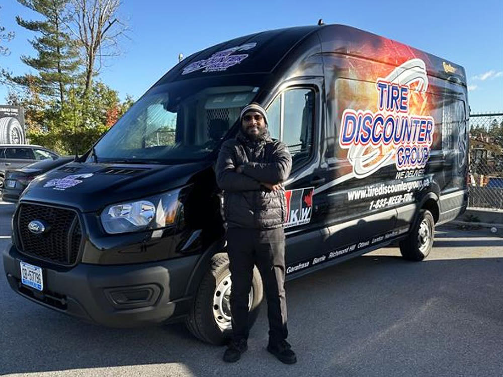 Conducteur en veste noire avec barbe, debout devant un camion Tire Discounter Group dans un parc de stationnement.