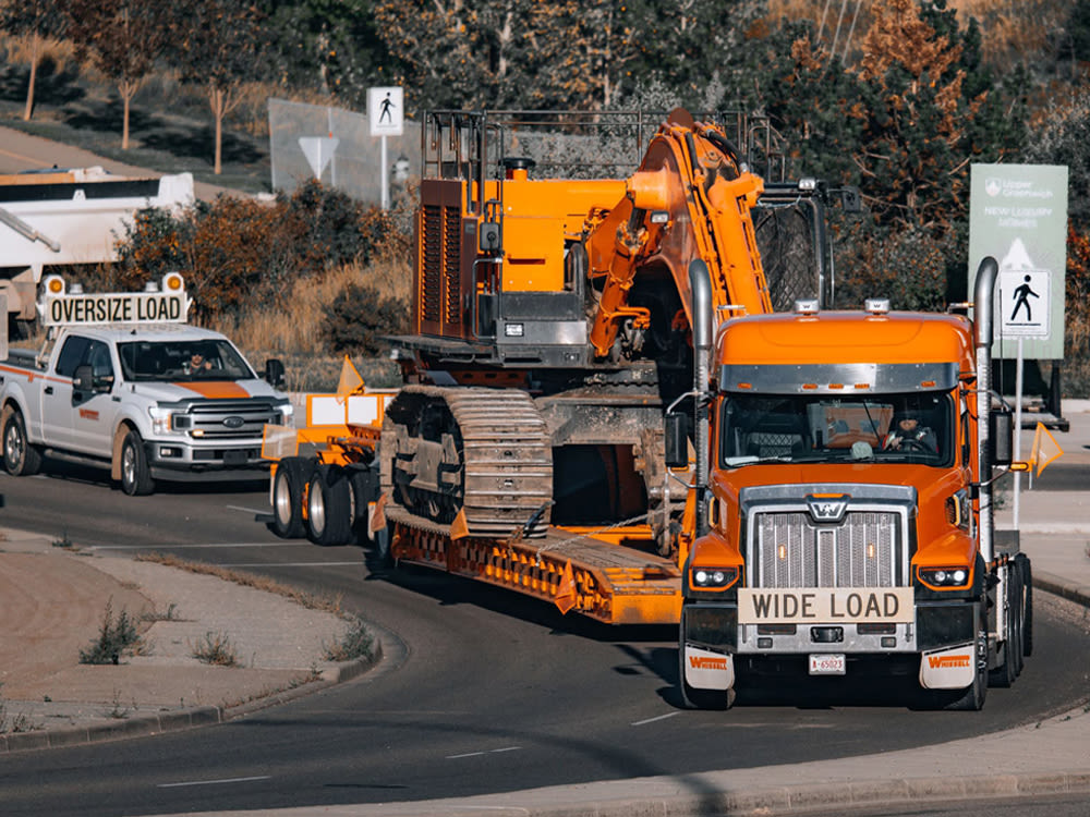Orange truck on road hauling excavator, followed by a white pickup truck with an oversize load placard