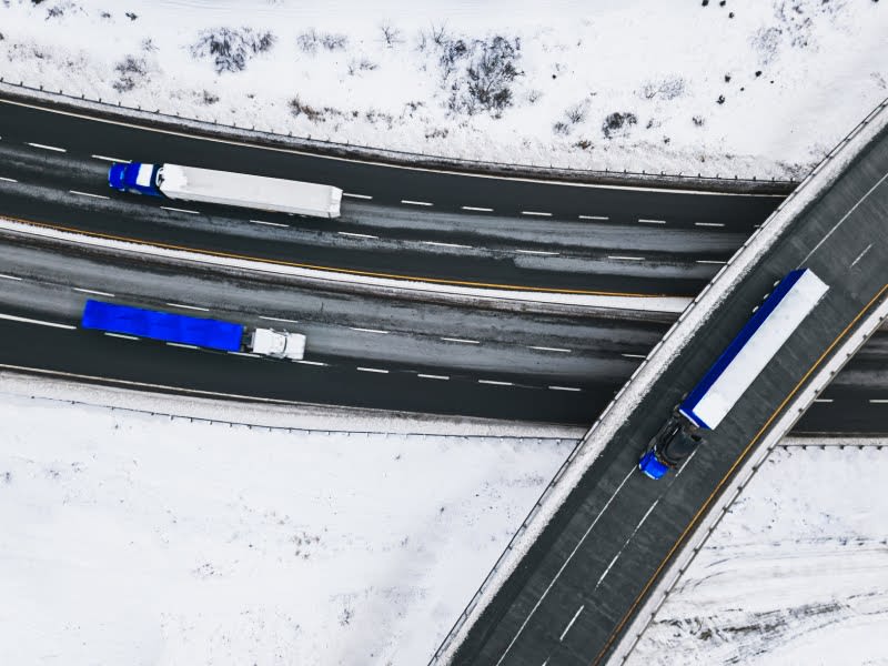 Aerial view of blue and white semi-trucks traveling on snowy highways with multiple road intersections.