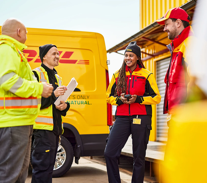 Two DHL employees looking at each other and smiling while standing in front of a DHL truck outside of a warehouse.