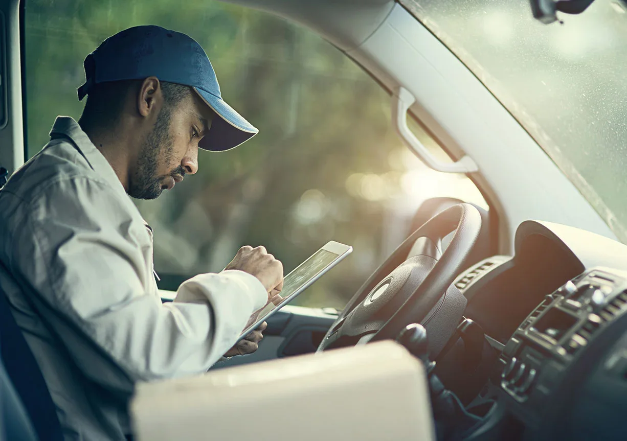Lieferfahrer mit blauer Kappe sitzt im Fahrzeug und bedient ein Tablet, ein Paket liegt neben ihm.