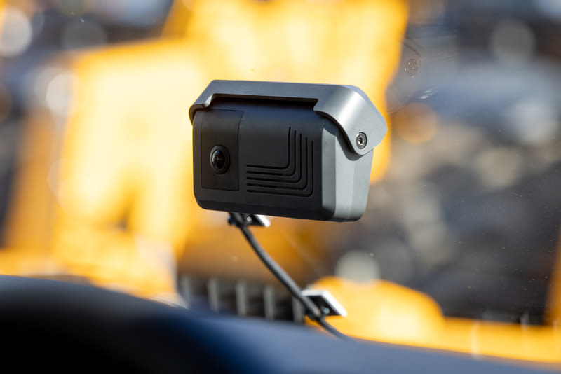 A small black and silver dash cam mounted on a vehicle windshield, with a yellow vehicle blurred in the background.