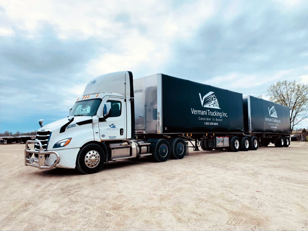 Semi-remorque Freightliner blanc avec deux remorques Vermani Trucking Inc. bleu foncé sur du gravier sous un ciel nuageux.