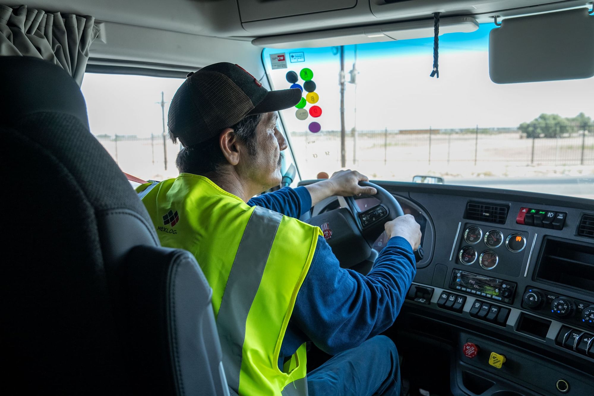 A truck driver in a yellow safety vest and black cap steers a large truck through a desert highway.