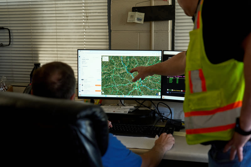 Two workers review a road map on a computer monitor, one in a yellow safety vest pointing at the screen.