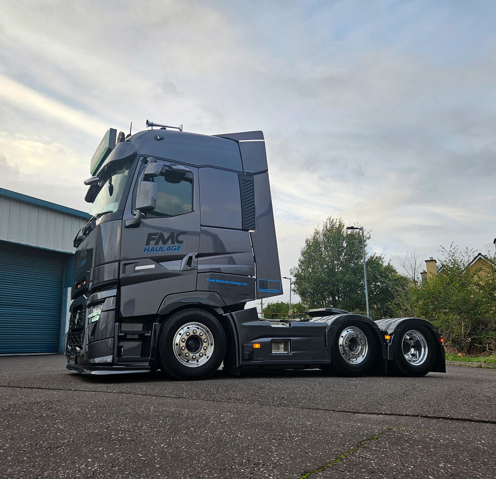 A dark grey FMC Haulage lorry cab parked in a yard, with chrome wheels, beside a blue industrial building.