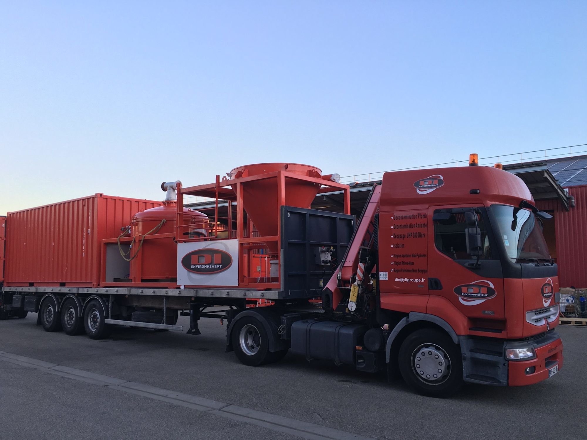 Camion rouge de transport industriel DLB Environnement avec équipement spécialisé sur remorque sous ciel bleu clair.