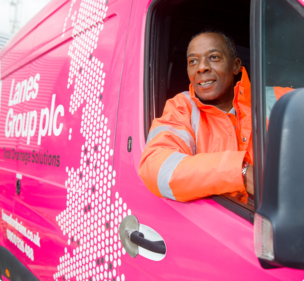 Chauffeur en veste orange fluorescente souriant depuis la fenêtre d'un camion rose de Lanes Group plc.