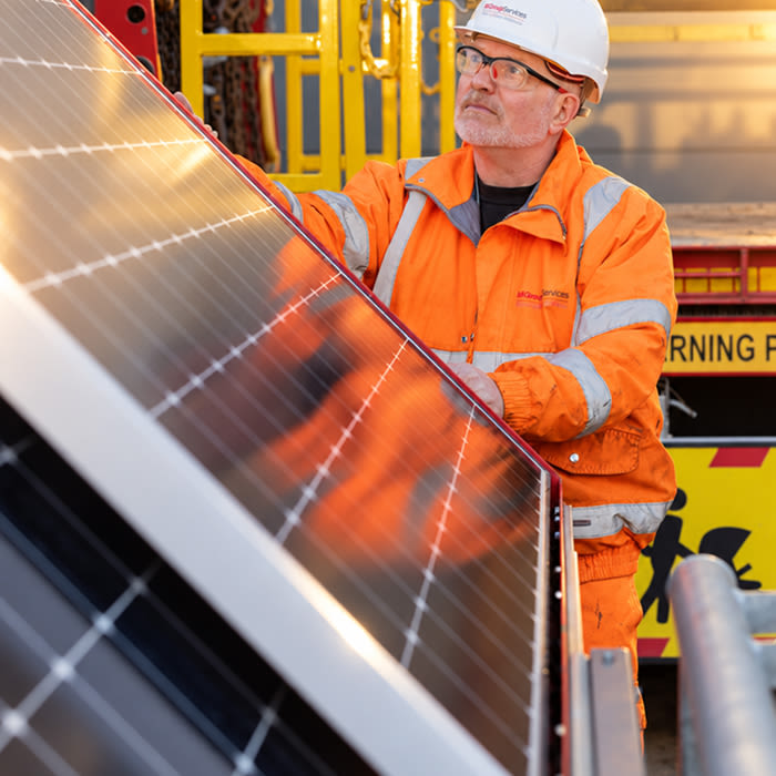 Worker in orange hi-vis jacket and white hard hat installing a solar panel outdoors.